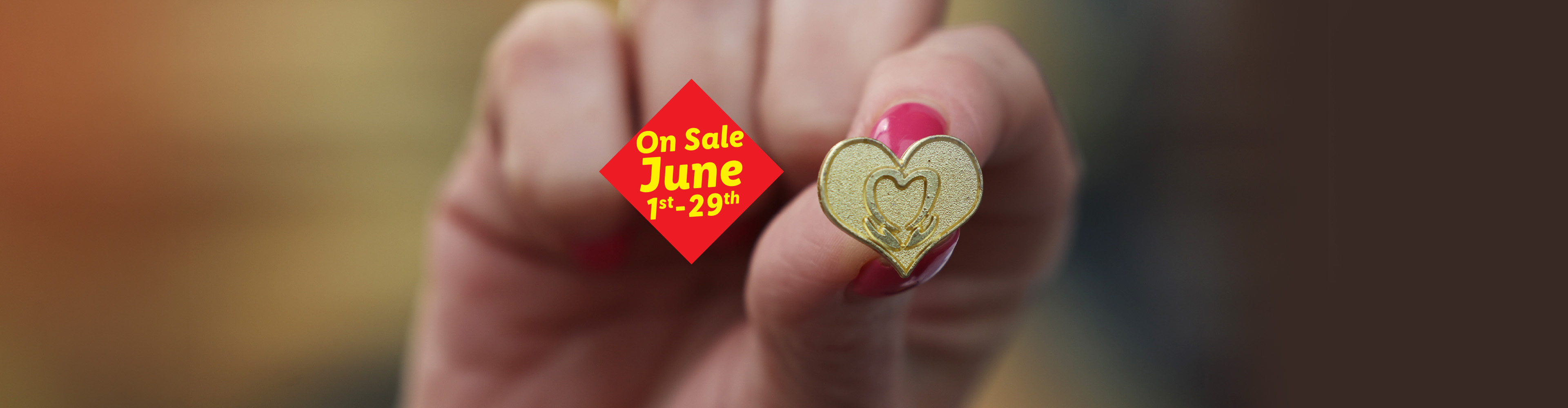 A hand holding a golden heart-shaped pin, with a red sale tag for June 1st-29th.