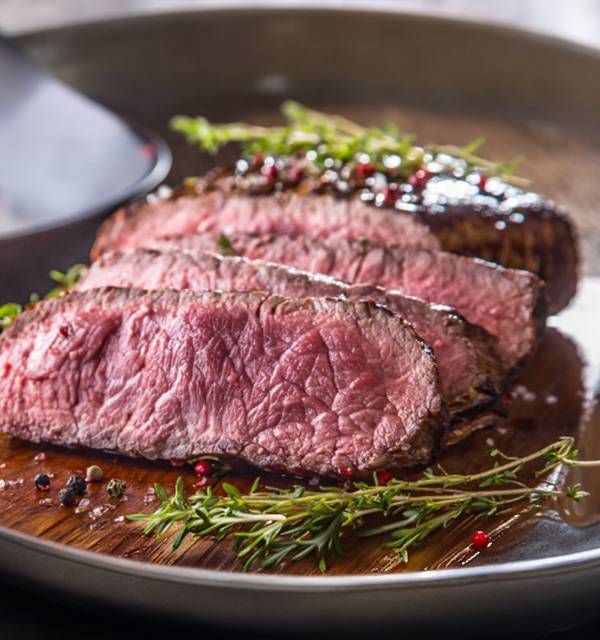Sliced medium-rare steak with herbs and peppercorns on a wooden board.