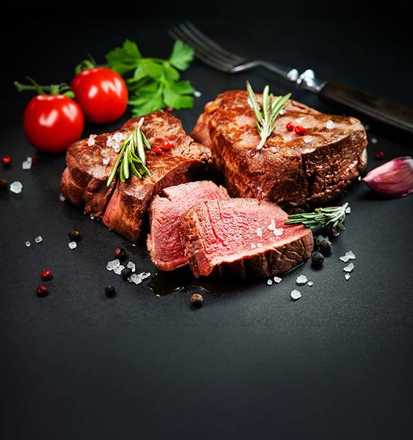 Cooked beef steaks with rosemary, tomatoes, and spices on a dark background.