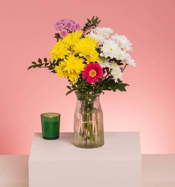 Vase of colorful chrysanthemums and a green candle on a white surface.