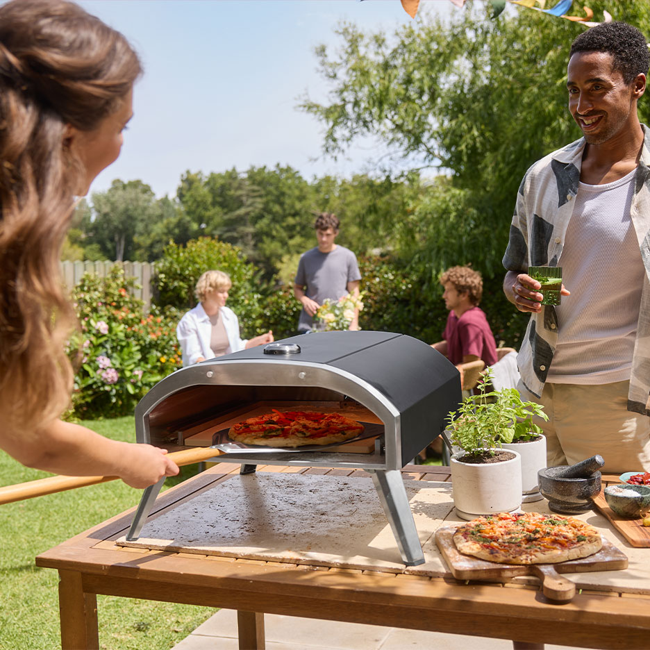 Family at a garden party, woman putting pizza into an oven, man holding a drink.